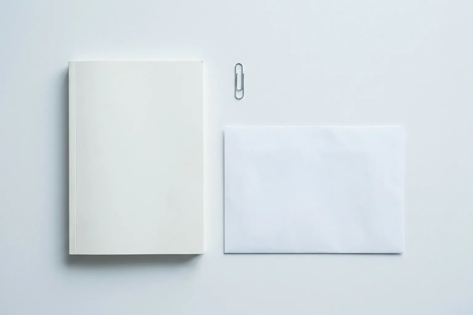 Dossier relié, trombone et grande enveloppe blanche disposés à plat sur un bureau blanc
